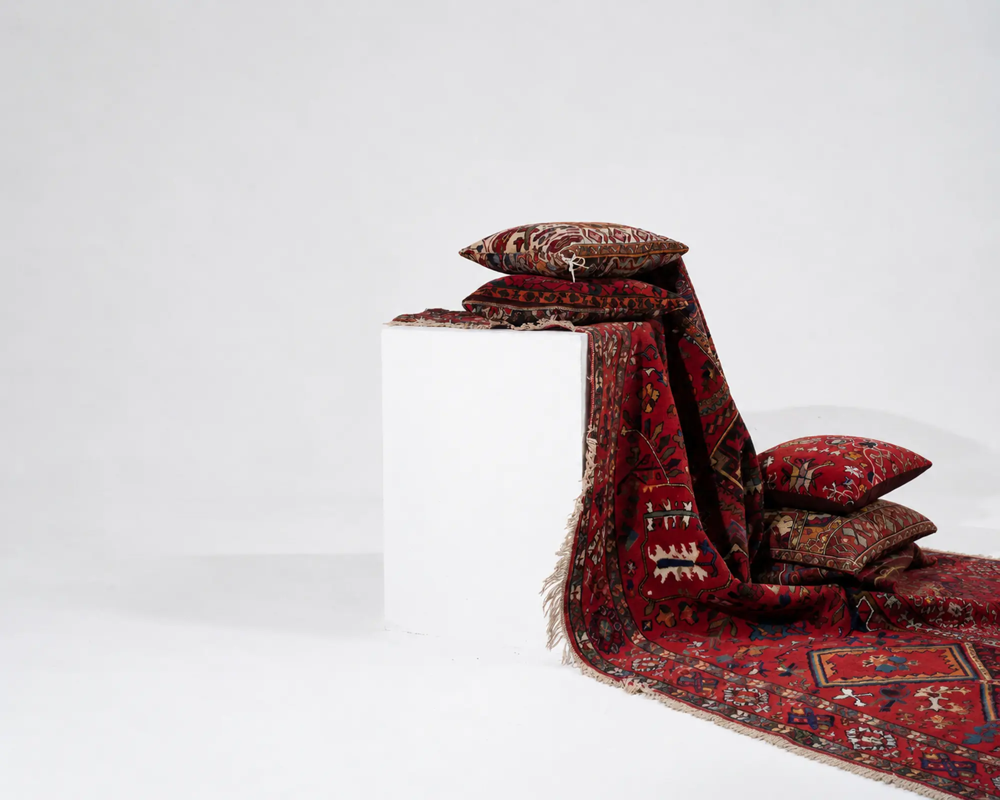 Handcrafted Persian cushions arranged with a red Persian rug on a white plinth in a minimalist studio.